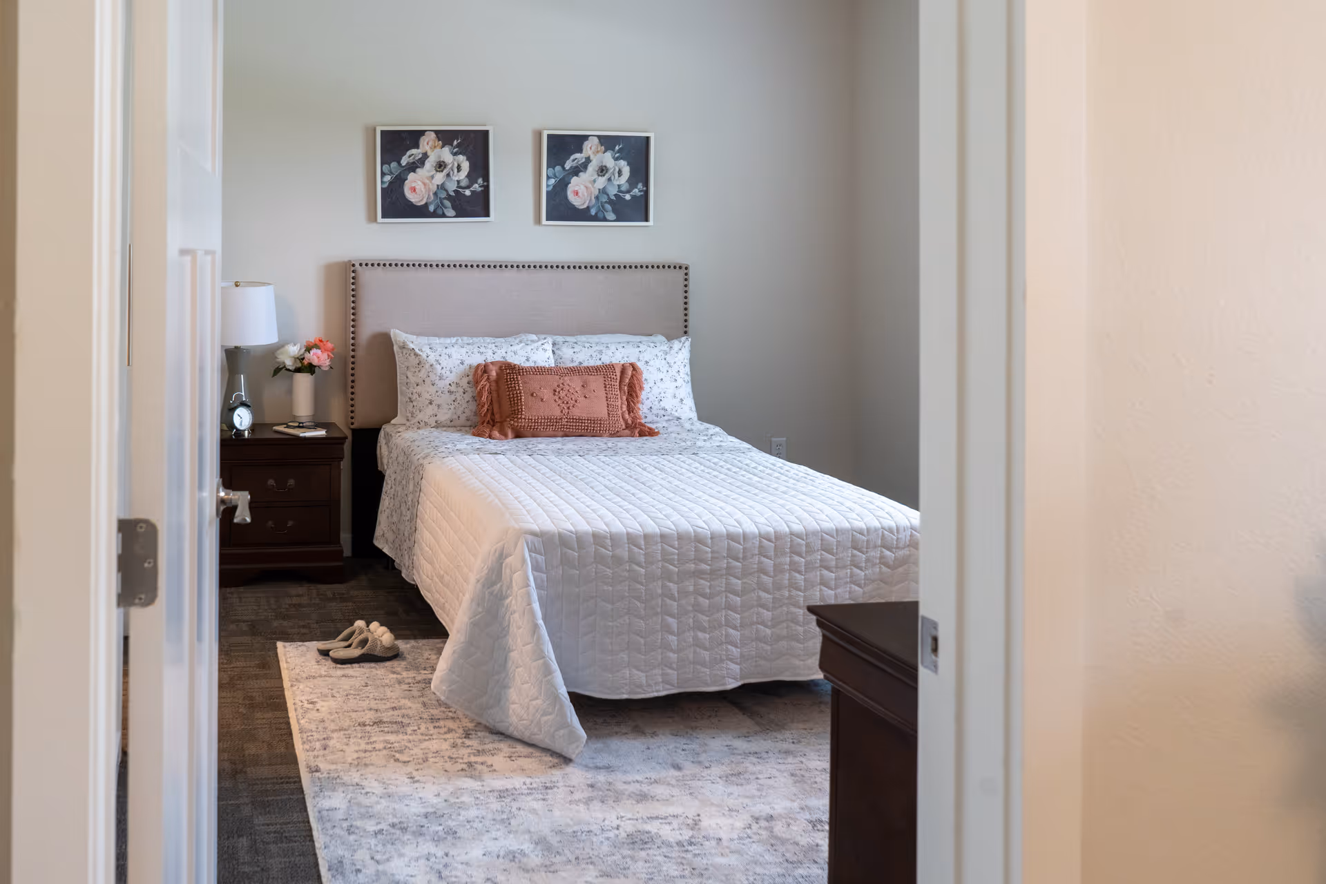 View through a doorway into a neat bedroom with a made bed, nightstand with a lamp and flowers, wall art, and a rug.