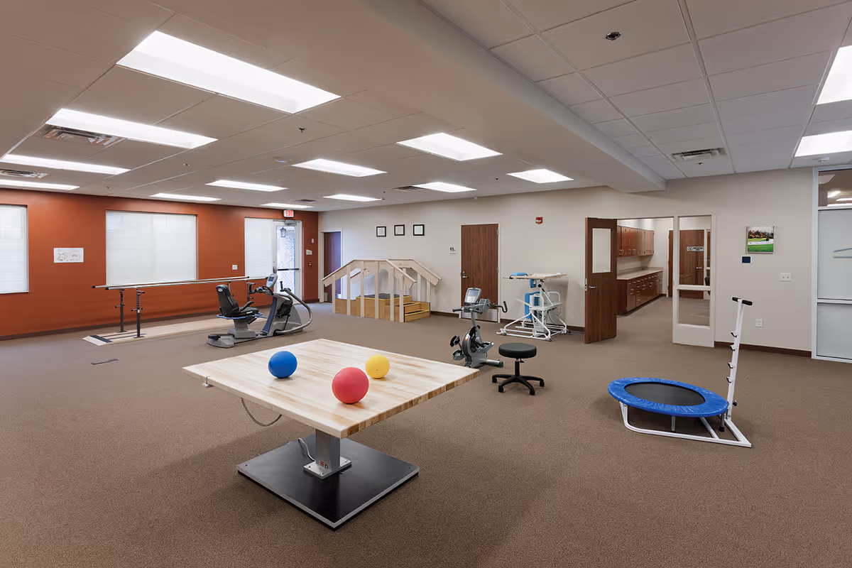 A spacious physical therapy or rehabilitation room with exercise equipment including stationary bikes, a small trampoline, parallel bars, a wooden table with colorful therapy balls, and a small set of stairs with handrails. The room has beige carpet, white ceiling tiles with fluorescent lighting, and walls painted in white and burnt orange. There are doors and windows along the walls, and a hallway leading to other rooms.