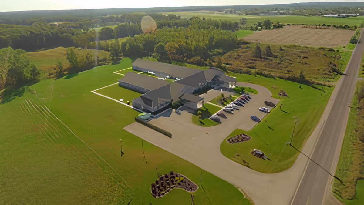 Aerial view of a senior living facility named Quality Care of Howell surrounded by green fields and trees, with a parking lot containing several cars and a road running alongside the property.