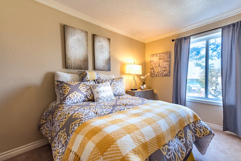 A cozy bedroom with a bed covered in patterned bedding and a yellow and white checkered blanket. There are decorative pillows including one with the word 'SNOOZE'. On the wall above the bed are two metallic sunburst wall art pieces. A nightstand with a lamp, a small clock, and a flower vase is next to the bed. A window with gray curtains lets in natural light and shows trees outside.