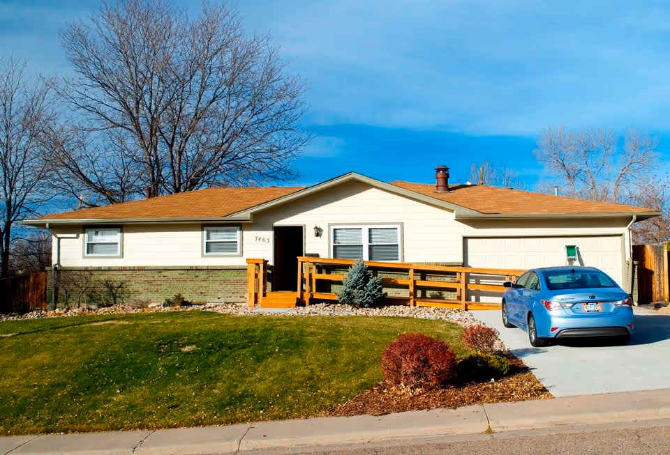 Single-story residential building with a brown roof, beige siding, and green brick lower exterior. There is a wooden ramp leading to the front door and a blue car parked in the driveway. Leafless trees are visible in the background under a clear blue sky.