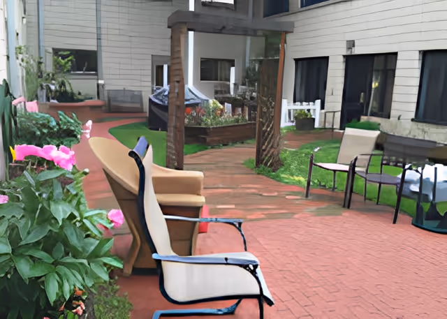 Outdoor patio area at Belle Reve Senior Living with several chairs arranged on a brick-paved surface, surrounded by green plants and flowers, adjacent to the building's exterior walls and windows.