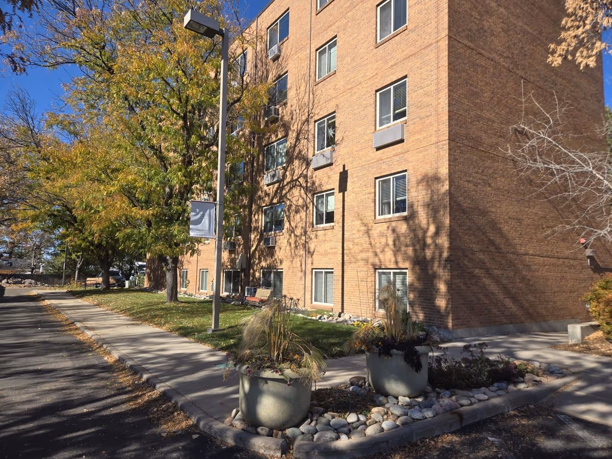 Brick multi-story building exterior with a sidewalk, large planters, trees, and a lamppost.