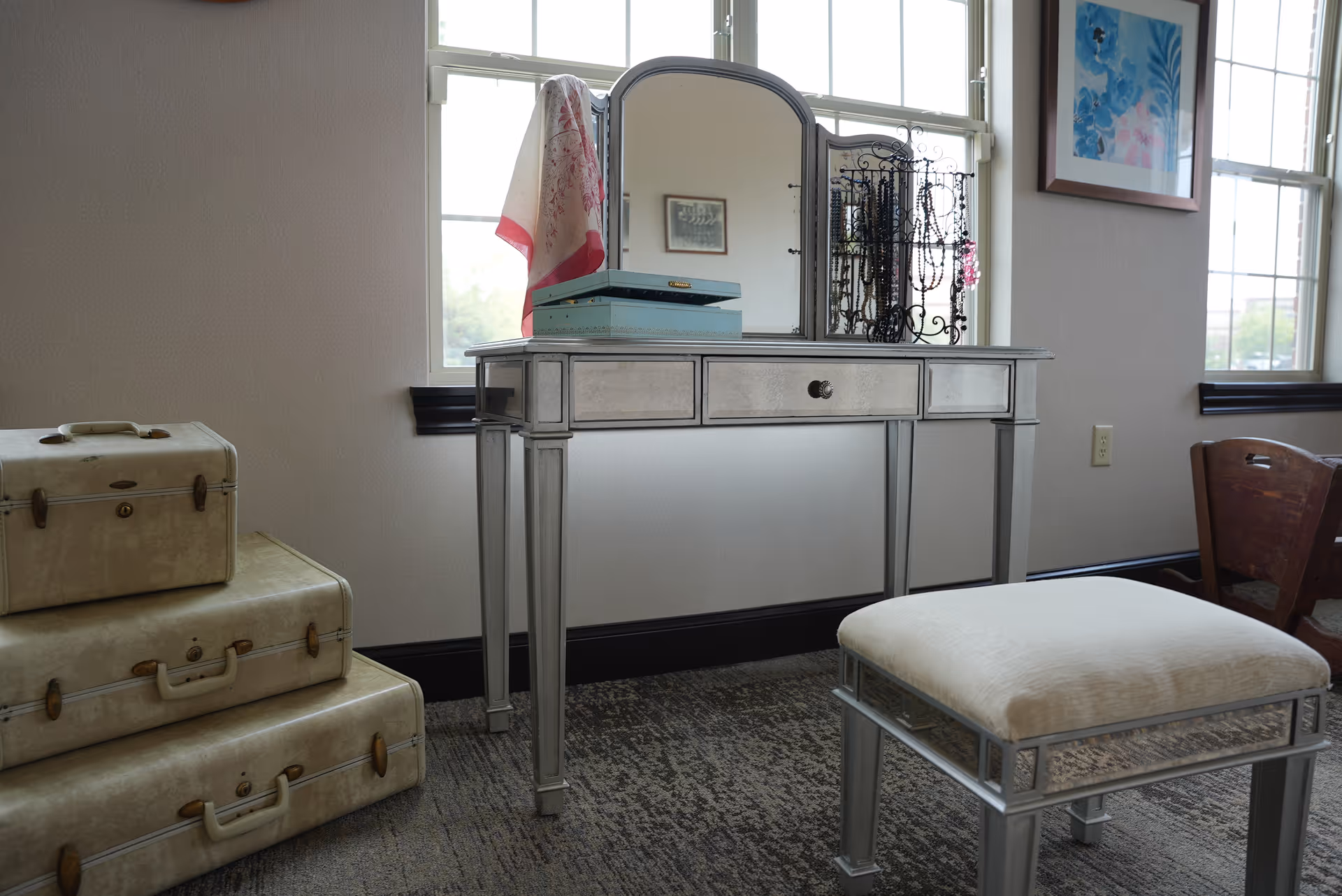 Vanity table with mirror, cushioned stool, and stacked suitcases beside windows in a furnished room.