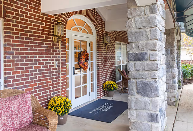Covered entrance area of a senior living facility with a brick wall and stone pillars. There is a white door with glass panels decorated with a fall wreath, two yellow potted flowers on either side of the door, and a black welcome mat. Wicker chairs with cushions are placed near the entrance.