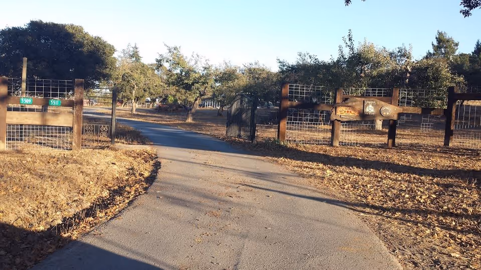 A paved driveway leading through a gated entrance with wooden and wire fencing on both sides. Trees and dry leaves are visible around the area under a clear sky.