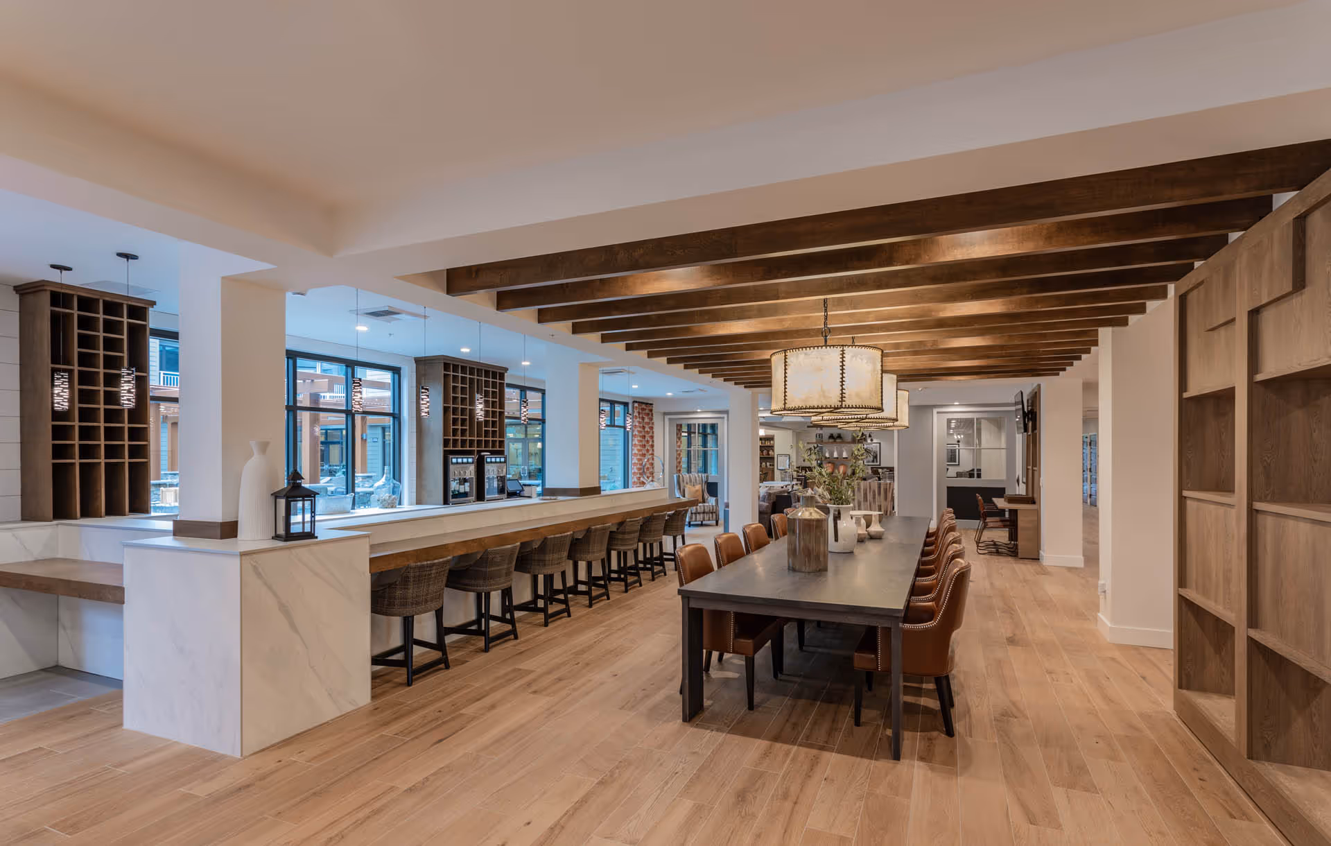 A spacious and modern dining area with a long wooden table surrounded by leather chairs. The ceiling features exposed wooden beams and large pendant lights. To the left, there is a long counter with bar stools and built-in wine racks, with large windows letting in natural light. The floor is light wood, and the overall decor is warm and inviting.