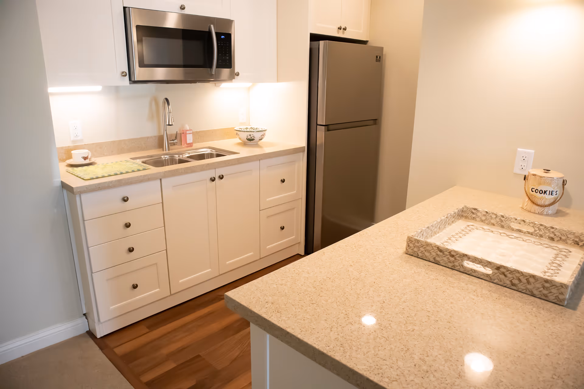 Small modern kitchenette with a sink and cabinets, microwave above, refrigerator, and a countertop island with a decorative tray.