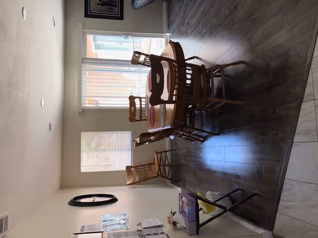 Round wooden dining table with several chairs in a bright room with large windows and hardwood floors.