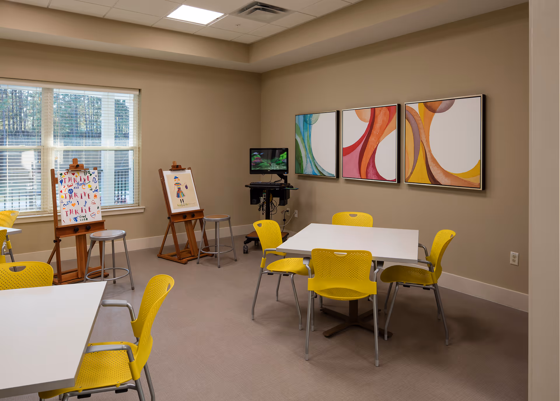 A bright activity room with beige walls and a large window letting in natural light. The room features two white tables surrounded by yellow chairs. There are two wooden easels displaying colorful artwork near the window, and a small TV mounted on a stand in the corner. Three abstract paintings with curved shapes in green, red, orange, and yellow tones hang on the wall.