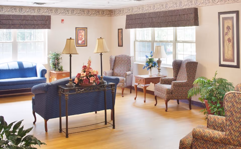 A bright and cozy living room area with large windows letting in natural light. The room features blue upholstered sofas, patterned wingback chairs, wooden side tables with lamps, floral arrangements, framed artwork on the walls, and several green plants. The floor is light wood, and the walls are light-colored with a decorative border near the ceiling.