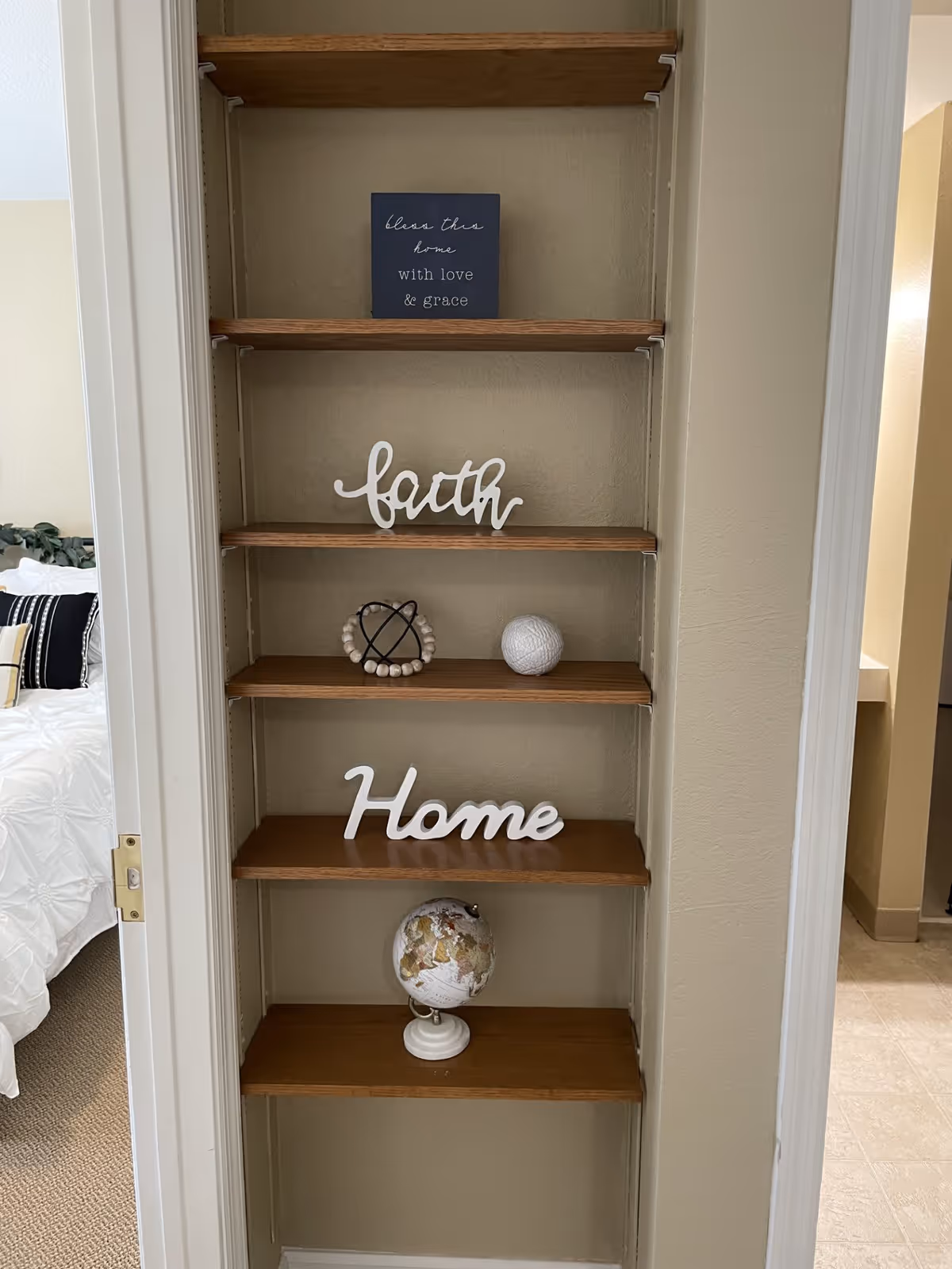 A built-in wooden shelf unit with five shelves displaying decorative items including a small sign that says 'bless this home with love & grace', a white 'faith' sign, a decorative orb, a white 'Home' sign, and a small globe. To the left, a bedroom with a white bedspread and pillows is partially visible. To the right, a hallway or another room with tiled flooring is visible.