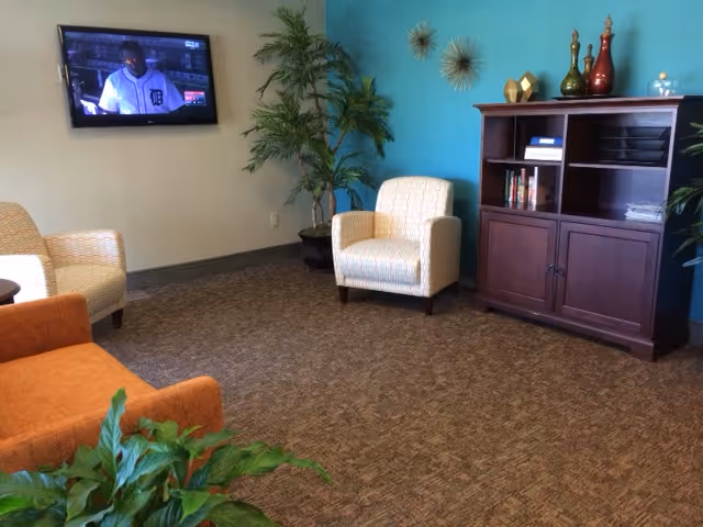 A cozy living room area with a wall-mounted TV showing a baseball game, two beige armchairs, an orange sofa, a wooden cabinet with decorative items on top, and green plants. The walls are painted light blue and beige, and the floor is carpeted.