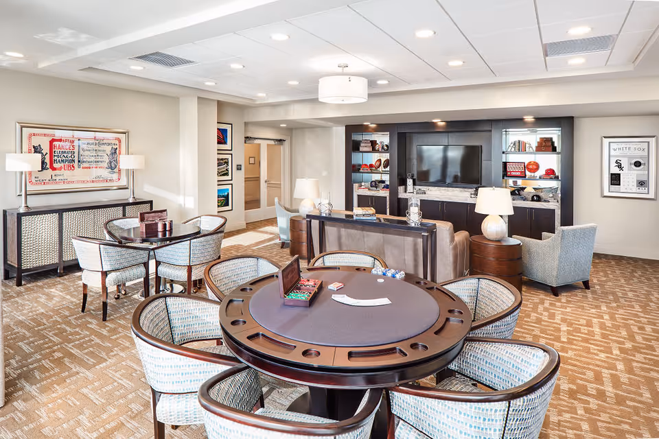 A bright and spacious common area in a senior living facility featuring a round poker table with six chairs in the foreground. Behind it, there is a seating area with a sofa, armchairs, side tables with lamps, and a large entertainment center with a flat-screen TV and shelves displaying decorative items. The room has carpeted floors, recessed lighting, and framed artwork on the walls.