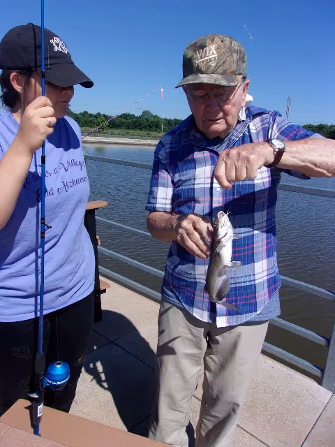 An elderly man wearing a camouflage cap and plaid shirt holds a fish he just caught while standing on a dock by the water. A woman wearing a black cap and a purple shirt with text on it stands next to him holding a fishing rod. The background shows a clear blue sky and a body of water with greenery on the far shore.
