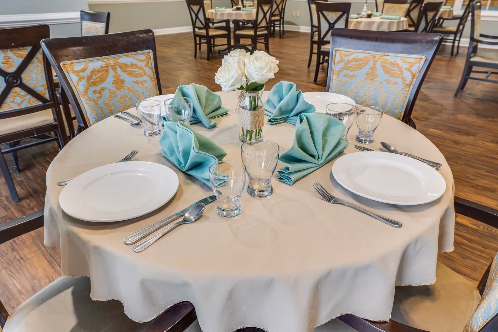 Round dining table set for four with plates, glasses, folded teal napkins and a white rose centerpiece in a communal dining room.