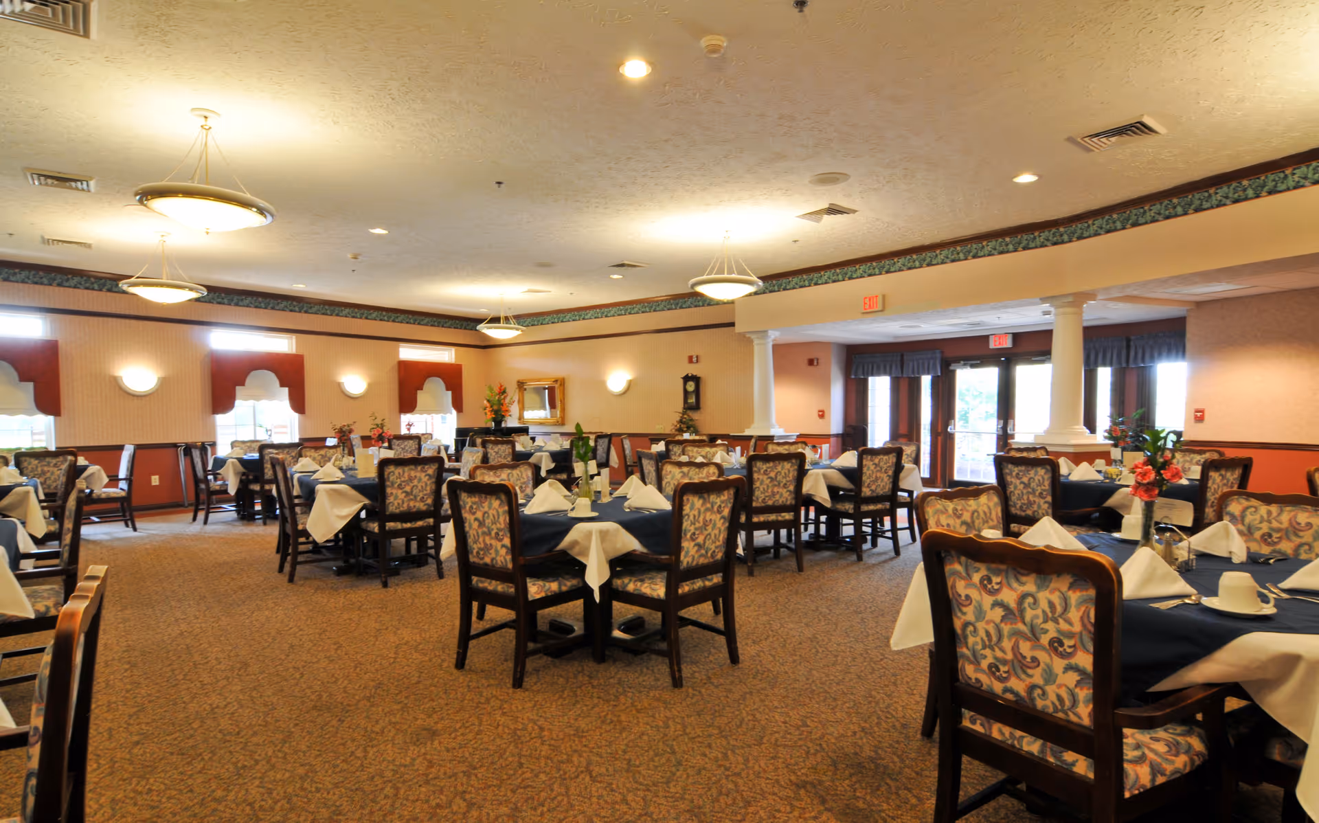 Spacious dining room with multiple set tables, patterned chairs, and ceiling lights in a senior living facility.