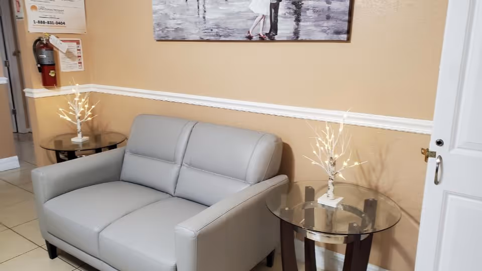 Small seating area with a gray loveseat between two glass side tables topped with decorative lighted branch lamps and a framed painting on the wall.
