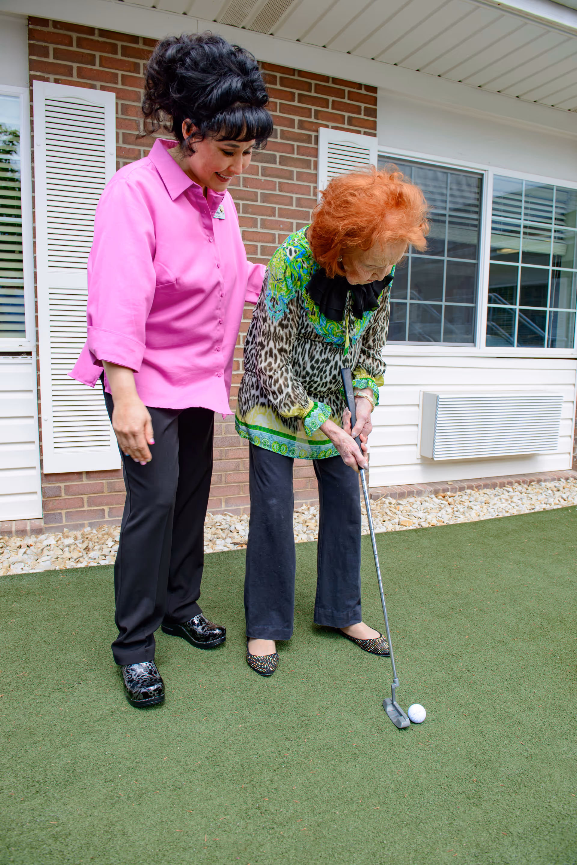 An elderly woman with red hair is playing mini golf on a green turf outside a brick building while a caregiver in a pink shirt stands beside her, smiling and offering support.