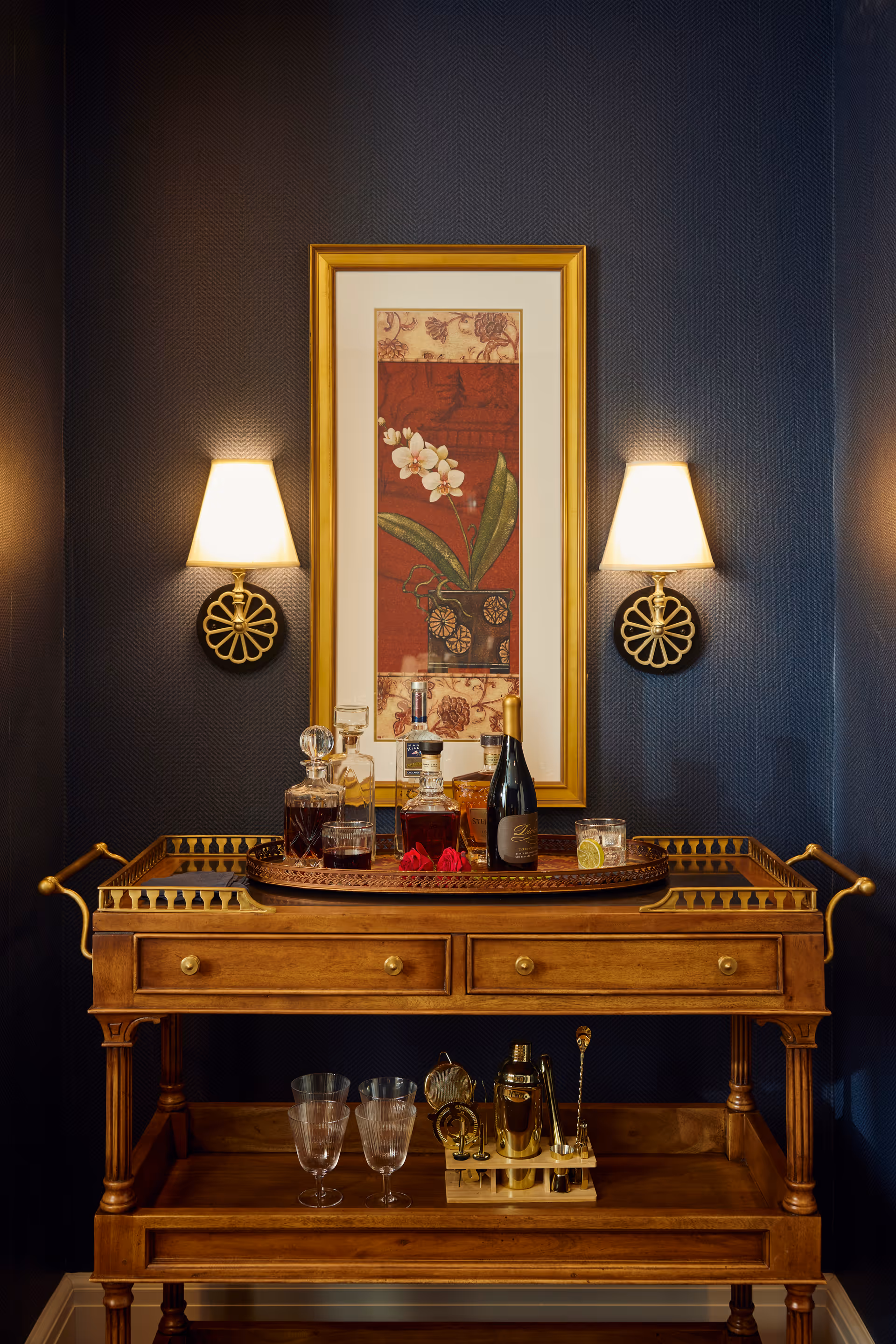 A wooden bar cart with two drawers and a lower shelf, holding various glass decanters, a bottle of wine, glasses, and a cocktail shaker set. The cart is positioned against a dark blue textured wall with two wall sconces on either side of a framed floral artwork featuring white flowers and green leaves on a red background.