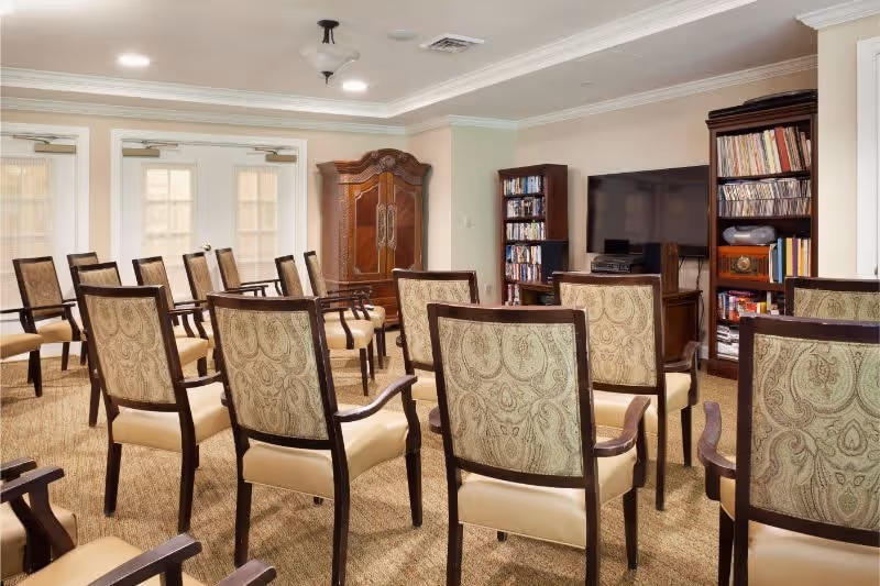 A room with multiple upholstered chairs arranged in rows facing a large flat-screen TV mounted on a wooden cabinet. There are bookshelves filled with books and DVDs on either side of the TV. The room has beige walls, carpeted floor, and double doors with glass panels at the back.
