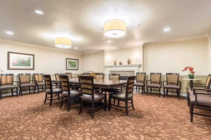 A well-lit communal dining room with a large central table surrounded by chairs, additional chairs lined along the walls, framed artwork, and a decorative fireplace.