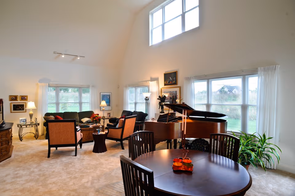 A spacious and well-lit living room with large windows and white curtains. The room features a grand piano, a round wooden dining table with chairs, a seating area with a sofa and armchairs, side tables with lamps, framed pictures on the walls, and indoor plants near the windows.