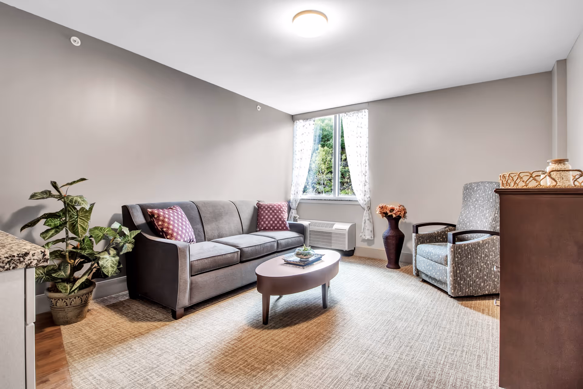 A cozy living room with a gray sofa adorned with two patterned pillows, a matching armchair, a small oval coffee table with a decorative centerpiece, a potted plant on the left, a tall vase with flowers near the window, and a window with sheer white curtains letting in natural light.