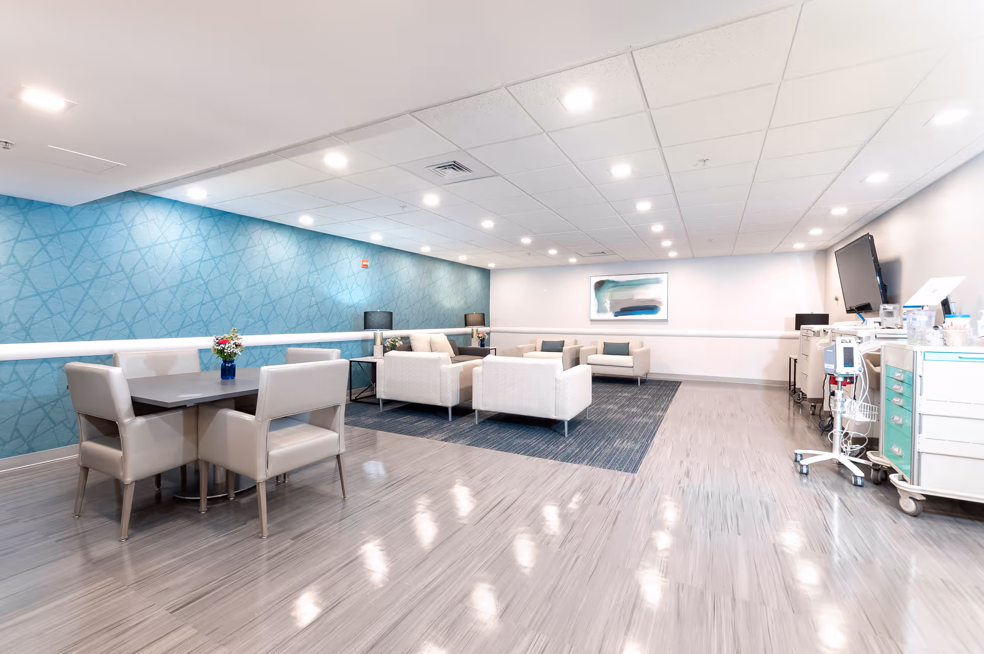 Bright common lounge with seating clusters, a dining table, blue accent wall and medical carts on the right.
