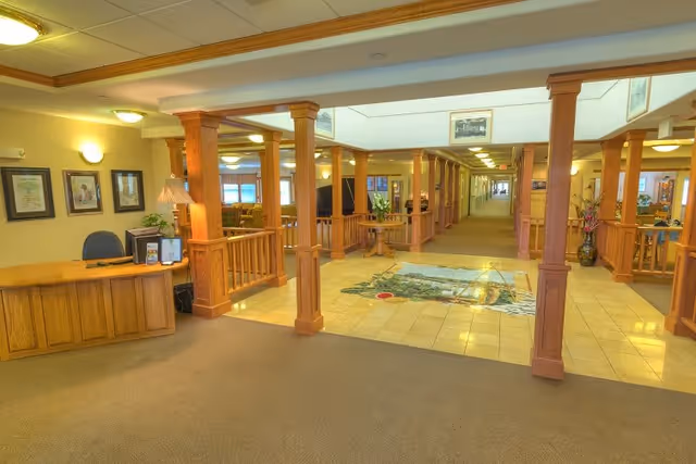 Interior view of a senior living facility lobby area with wooden pillars and railings, a reception desk on the left with a chair and framed pictures on the wall, a tiled floor with a decorative inlay in the center, and a long hallway extending into the distance with seating areas visible on the sides.