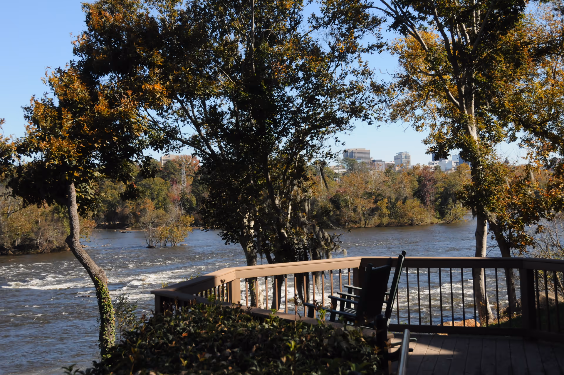 Wooden riverside deck with rocking chairs overlooking a tree-lined river and distant city skyline.