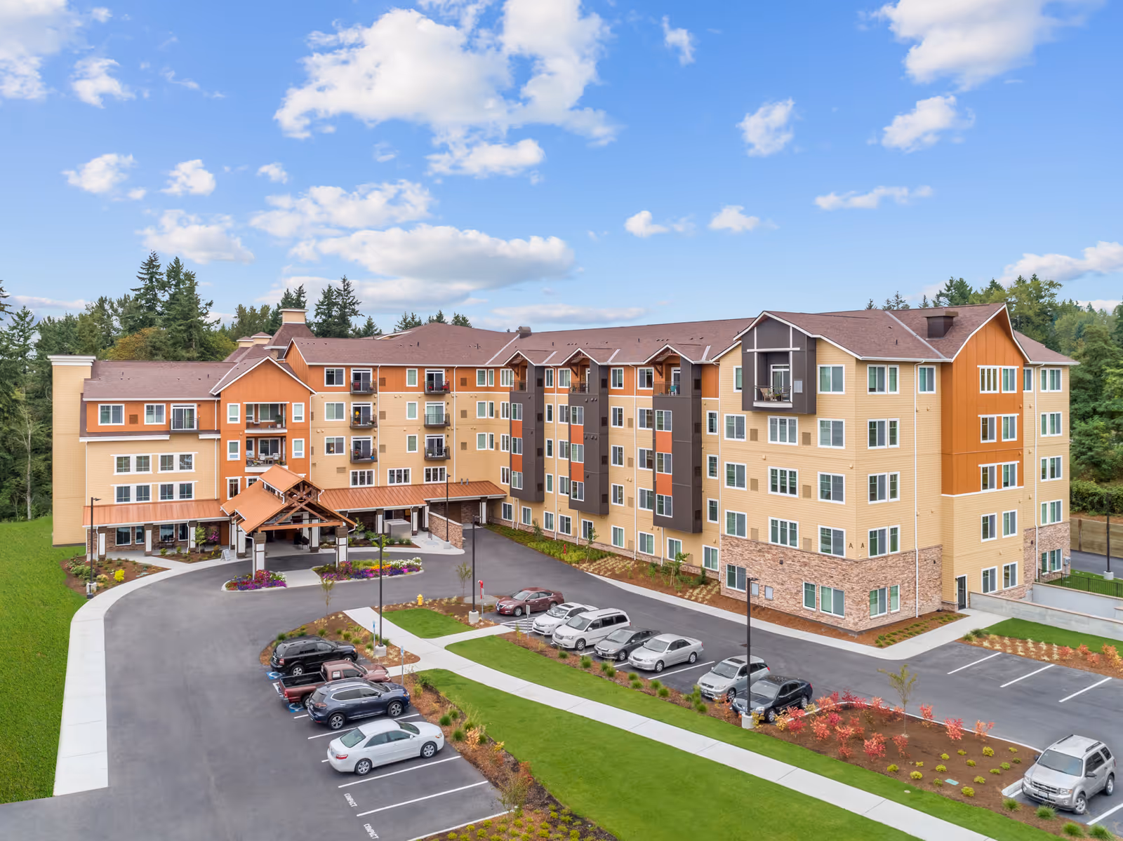Exterior view of a large, multi-story senior living facility building with a covered entrance, surrounded by a parking lot and landscaped greenery under a partly cloudy sky.