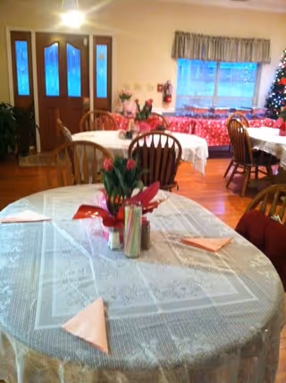 A dining area with round tables covered in white lace tablecloths, each set with folded napkins and a centerpiece of flowers. Wooden chairs surround the tables. In the background, there is a window with a valance, a decorated Christmas tree, and a door with glass panels. The floor is wooden.