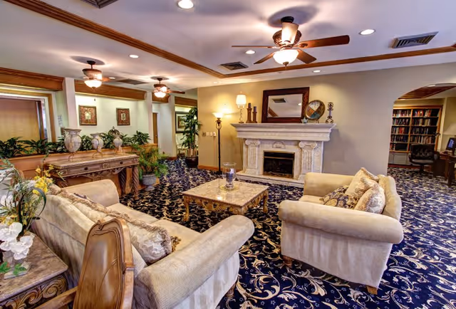 A cozy living room area in a senior living facility featuring two beige upholstered sofas with decorative pillows, a marble coffee table, a fireplace with a mantel holding a mirror and decorative items, ceiling fans with lights, patterned blue carpet, and plants along the walls.