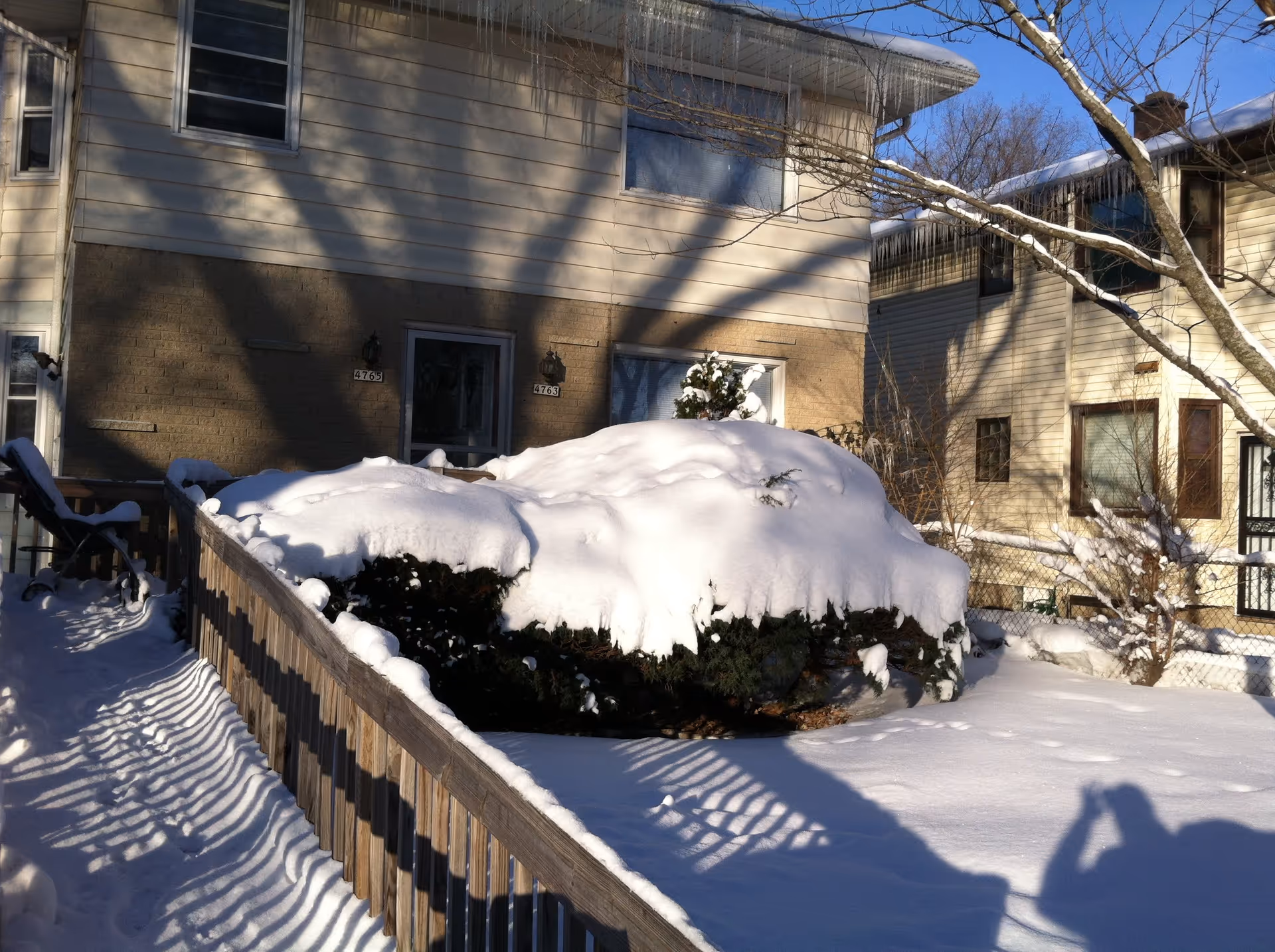 Snow-covered bushes and ground in front of a two-story residential building with beige siding and brick lower walls. A wooden railing runs along a snow-covered walkway leading to the building's entrance. Icicles hang from the roof edge, and leafless tree branches are visible.
