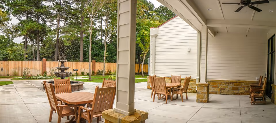 Outdoor patio area with wooden tables and chairs under a covered section next to a building. There is a multi-tiered water fountain on a concrete patio surrounded by a wooden fence and trees in the background.