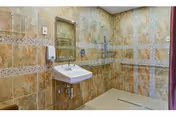 A spacious bathroom with beige and brown tiled walls and floor, featuring a wall-mounted sink with a mirror above it, a soap dispenser, and a walk-in shower area with a grab bar and a handheld showerhead.