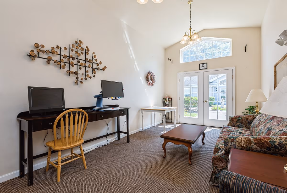 A bright living room with a floral patterned sofa, wooden coffee table, and a wooden desk with two computer monitors and a wooden chair. There is a decorative wall art piece above the desk, a small table near the glass double doors that lead outside, and a chandelier hanging from the ceiling.