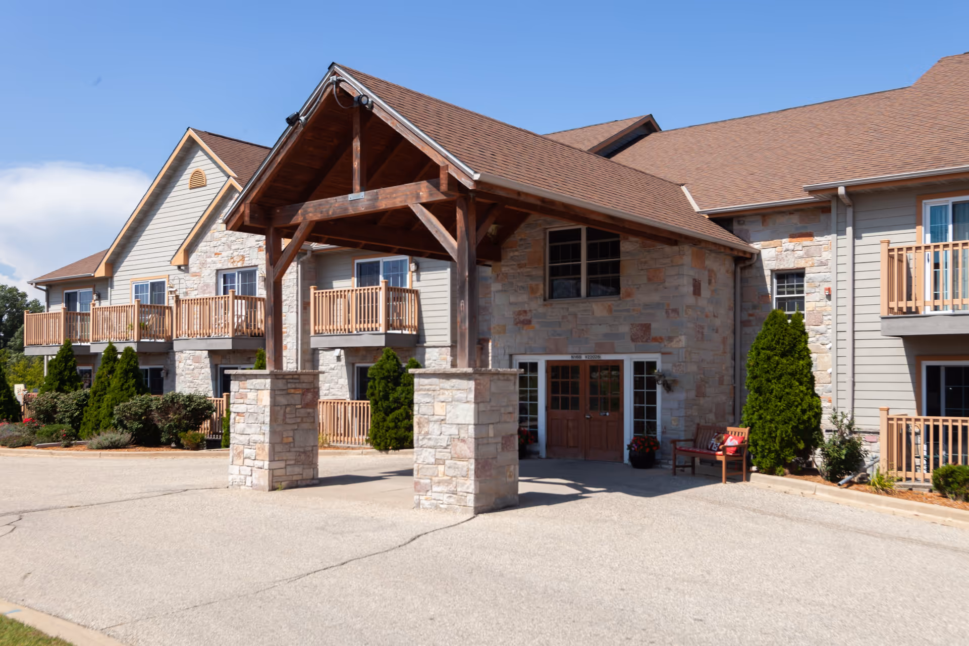 Exterior view of Charter Senior Living of Hasmer Lake showing a stone and wood building with a covered entrance supported by stone pillars. The building has balconies with wooden railings, greenery, and a clear blue sky in the background.