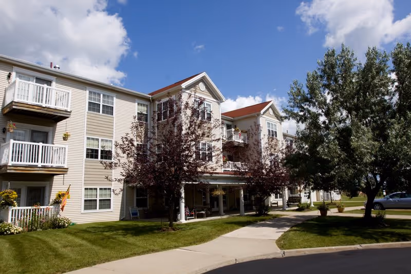 Exterior view of a multi-story residential building with balconies, surrounded by green lawns and trees under a partly cloudy blue sky.