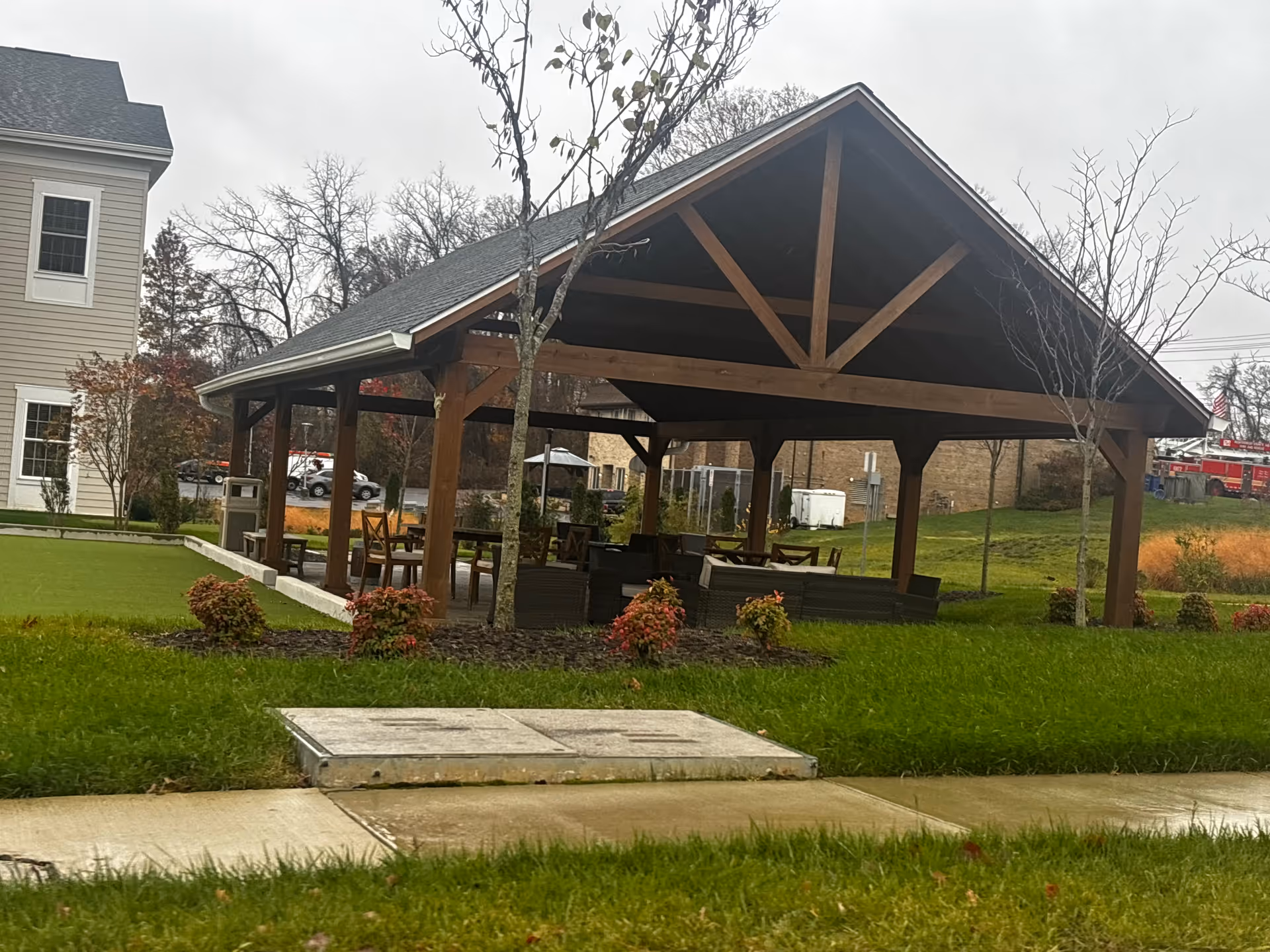Outdoor covered pavilion with wooden beams and roof, furnished with tables and chairs, surrounded by green grass and small shrubs, with a sidewalk in the foreground and a building partially visible on the left.
