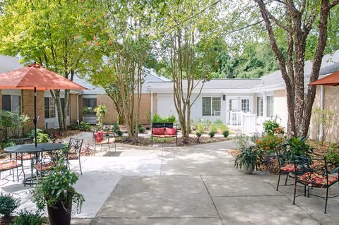 Sunny courtyard with patio tables, chairs, umbrellas, potted plants, trees, and low brick buildings surrounding it.