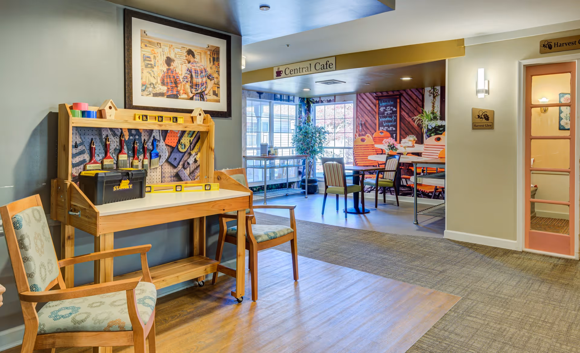 Interior view of a senior living facility showing a small workshop area with a wooden workbench holding paintbrushes, a toolbox, and measuring tools. Two wooden chairs with patterned cushions are placed nearby. In the background, there is a dining area with tables and chairs, large windows letting in natural light, and signs reading 'Central Cafe' and 'Harvest Glen'.