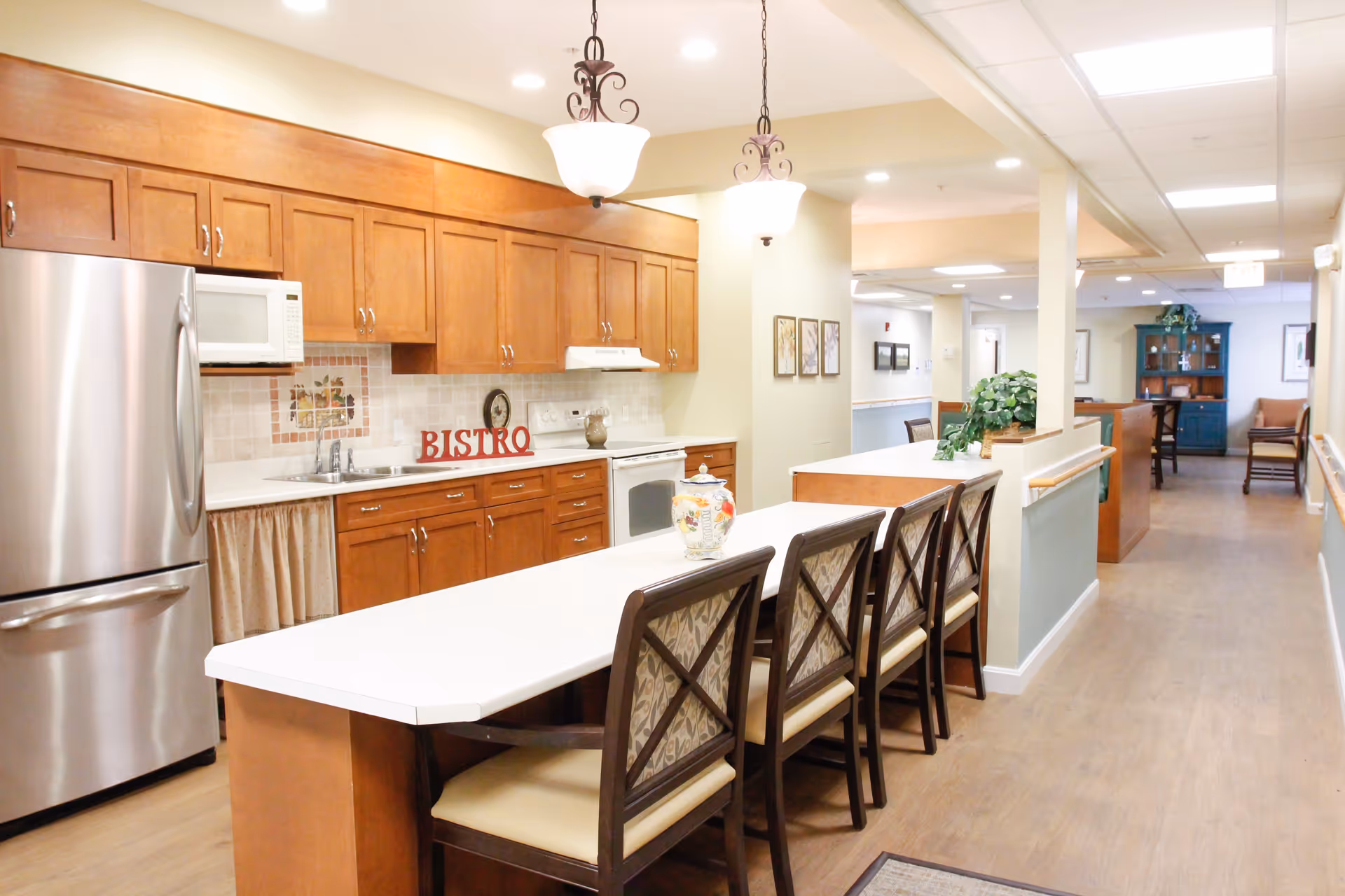 A bright and clean kitchen area in a senior living facility featuring wooden cabinets, a stainless steel refrigerator, a microwave, a stove, and a long white countertop with four cushioned chairs. The word 'BISTRO' is displayed on the backsplash above the sink. The kitchen opens into a hallway with additional seating and decorative plants.