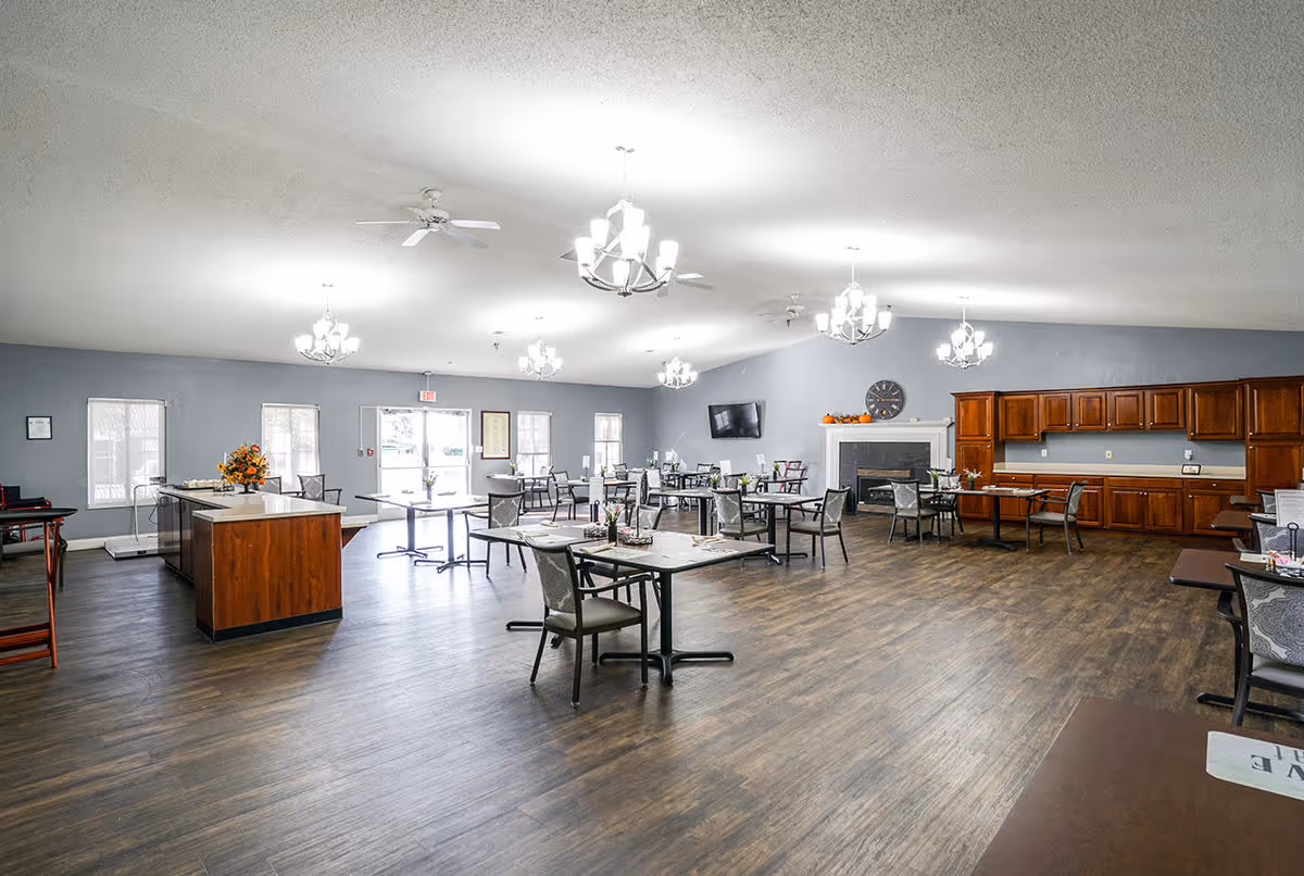 Spacious communal dining room with multiple tables and chairs, a fireplace, wall-mounted TV, and built-in cabinets under chandelier lighting.