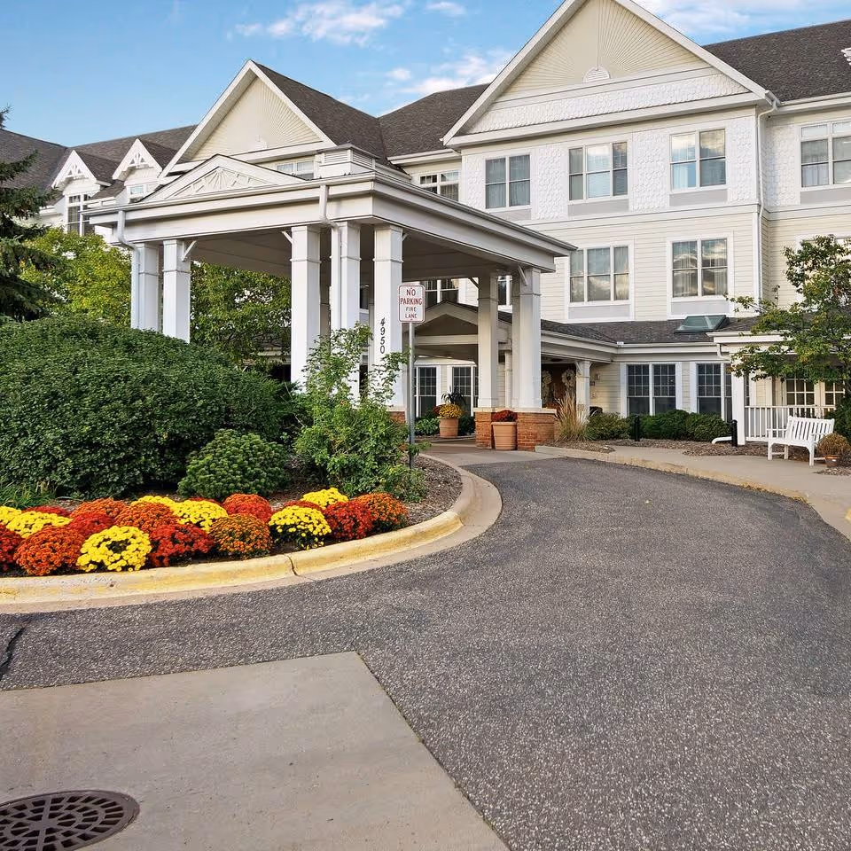 Entrance of a senior living facility with a covered drop-off area, surrounded by well-maintained landscaping including colorful flowers and green bushes. The building is white with multiple windows and a peaked roof under a blue sky with some clouds.