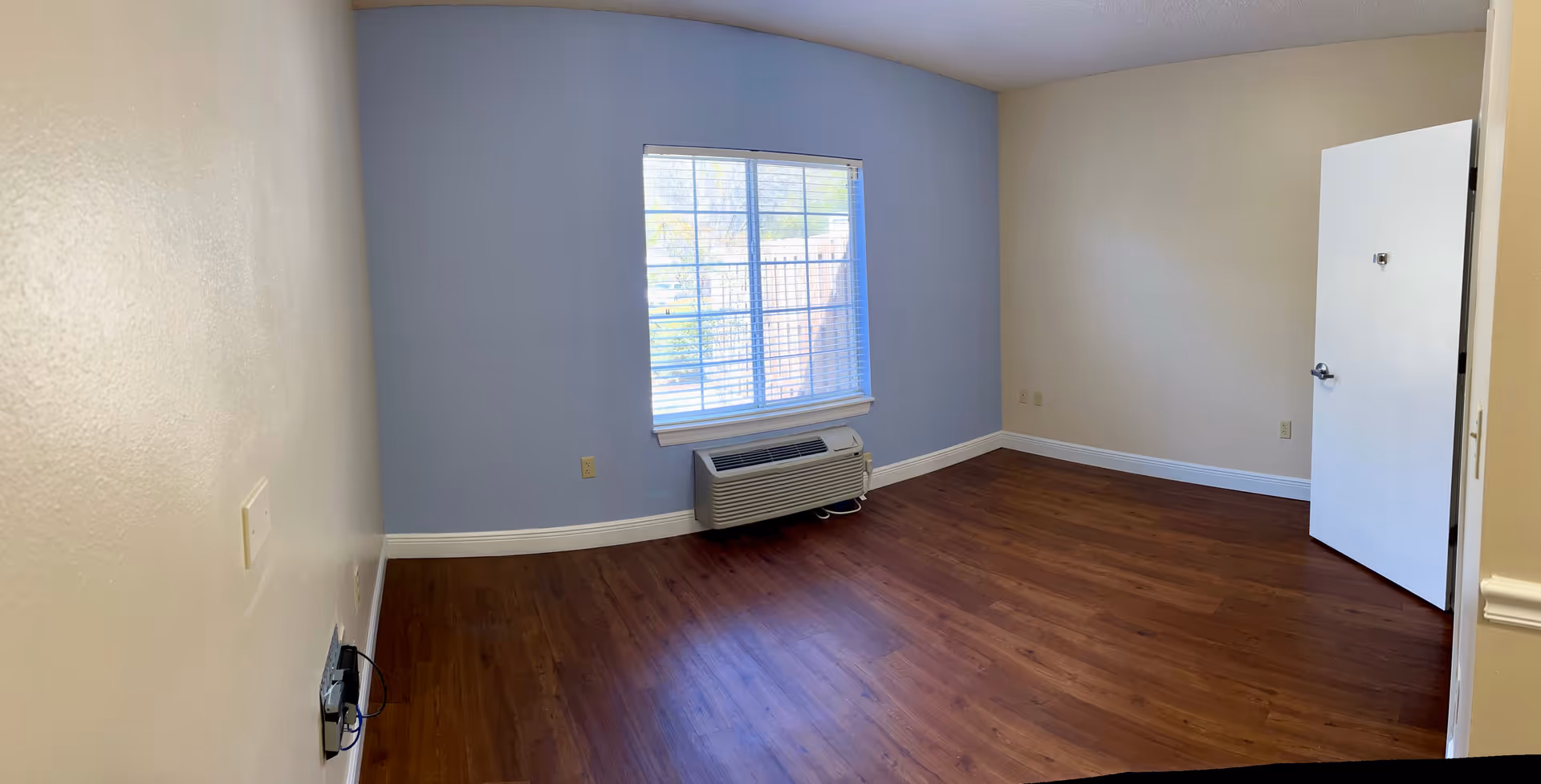 Empty senior living bedroom with wood floors, a window with blinds and an HVAC unit beneath it, and an open door.