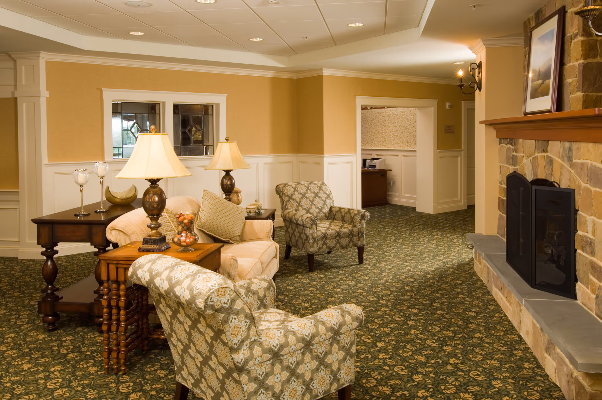 Cozy senior living common room with upholstered armchairs, side tables and lamps, patterned carpet, and a stone fireplace.