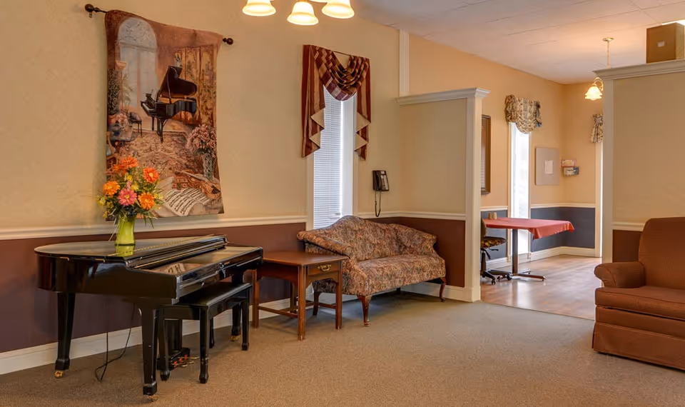 A cozy interior room featuring a black grand piano with a vase of colorful flowers on top, a floral patterned loveseat, a wooden side table, and an upholstered armchair. The walls are decorated with a tapestry depicting a piano and a window with draped curtains. In the background, there is a small table with a red tablecloth and chairs near windows letting in natural light.