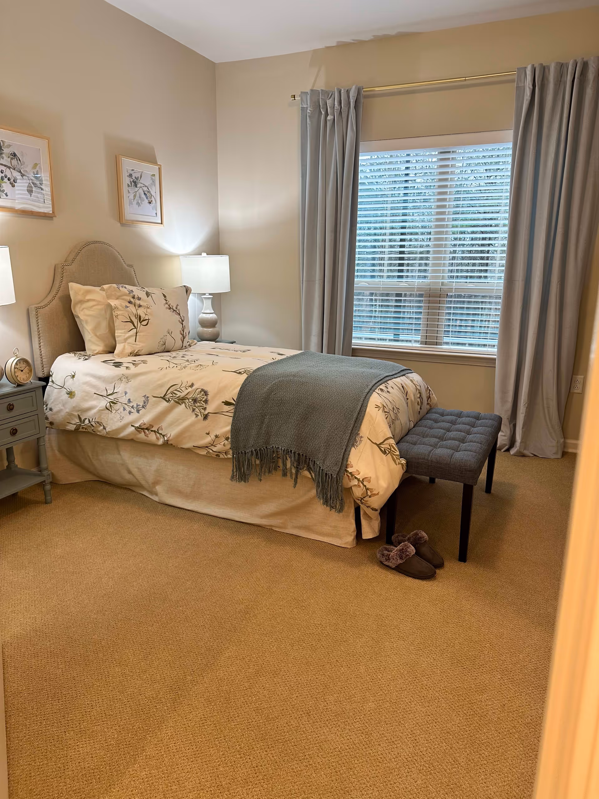 Cozy bedroom featuring a bed with floral bedding, a nightstand with a lamp, and a window with light curtains.